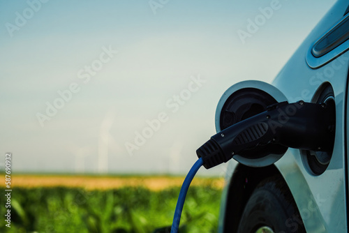 Electric automobile charges via EV plug against blurred windmills under blue sky. Wind turbines generate renewable energy in countryside