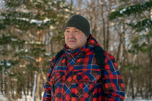 A man in a red plaid jacket and a gray hat walks in a pine forest