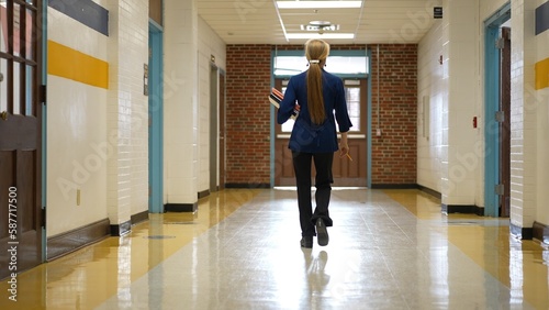Rear view of teacher walking down a hallway in an empty school holding books showing emptiness.