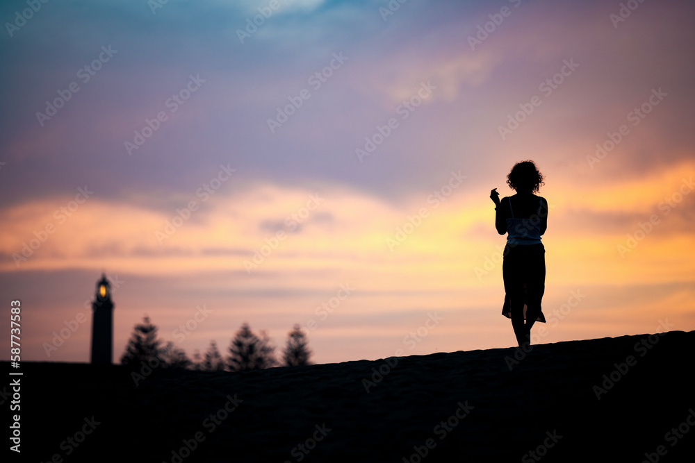 Stunning view of a silhouette of a person walking on some sand dunes at sunset with the Maspalomas lighthouse in the distance. Gran Canaria, Canary Islands