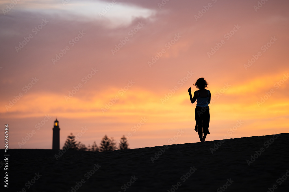 Stunning view of a silhouette of a person walking on some sand dunes at sunset with the Maspalomas lighthouse in the distance. Gran Canaria, Canary Islands