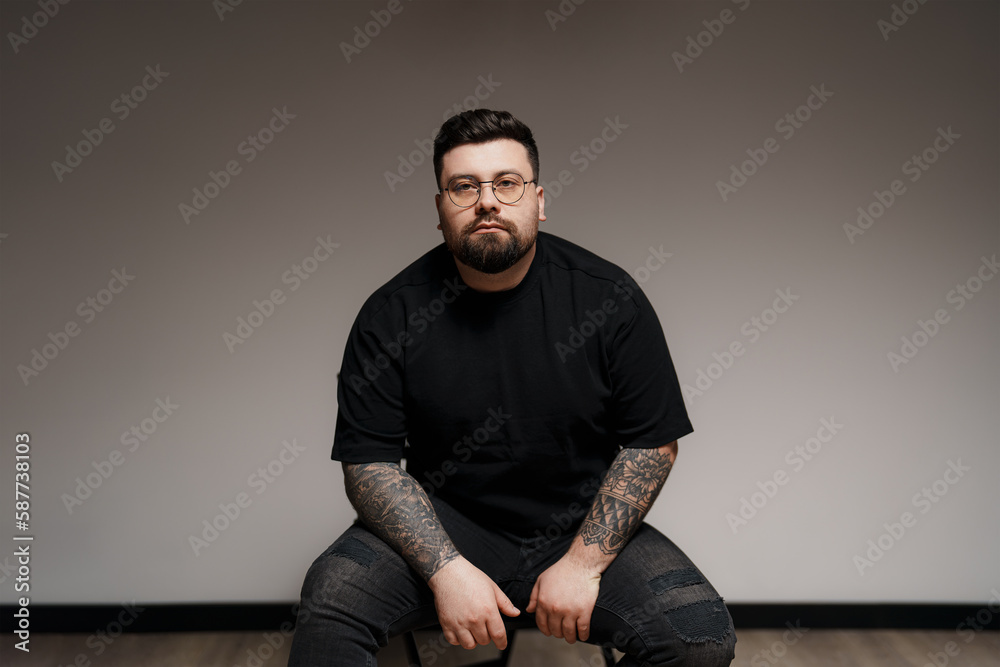 Content young plus size bearded male model in black outfit and eyeglasses with tattooed arms sitting on chair against gray background in studio Content young plus size bearded male model in black outfit and eyeglasses with tattooed arms sitting on chair against gray background in studio