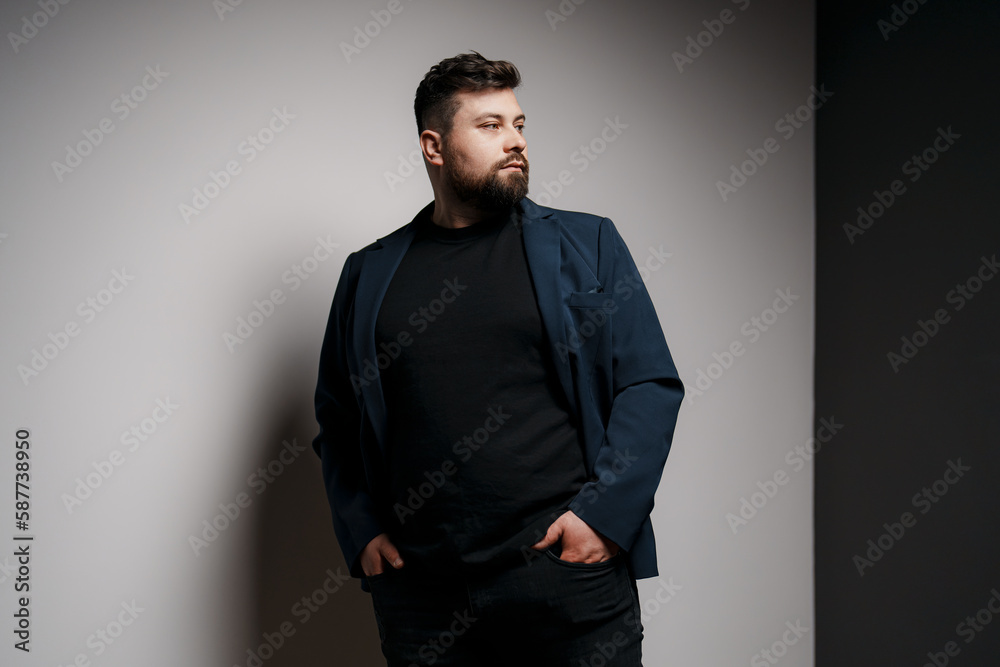 Fototapeta premium Pensive young bearded male model in elegant jacket and black pullover standing in studio with hands in pockets and looking away thoughtfully