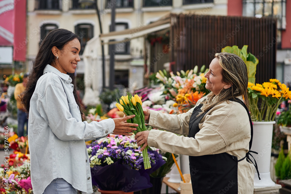 Fototapeta premium Young Latina woman purchasing vibrant flowers and plants from a street vendor's stall, handed by the gardener.