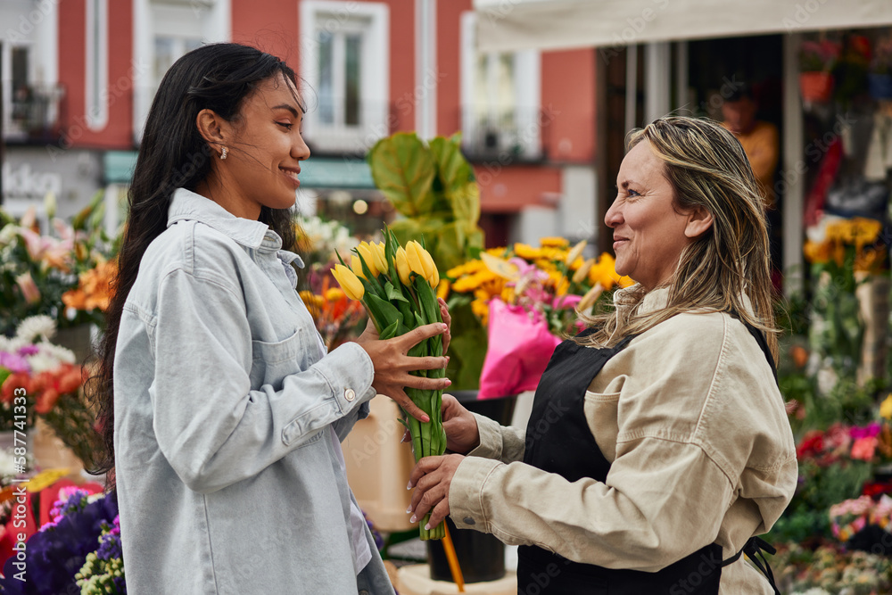 Young Latina woman purchasing vibrant flowers and plants from a street vendor's stall, handed by the gardener.