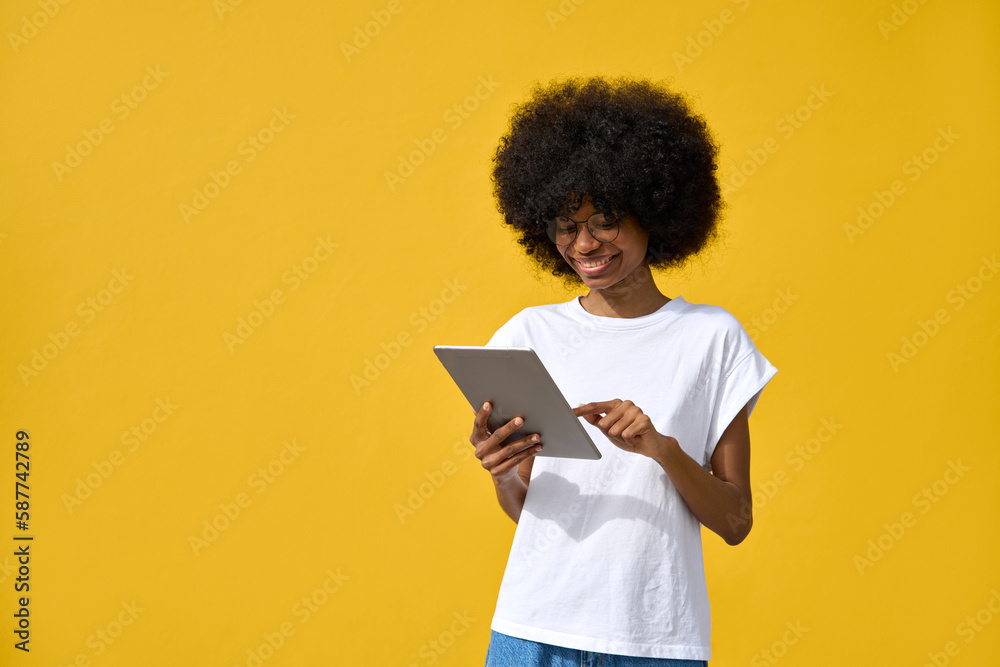 A happy hipster girl with afro haircut enjoy online communication and online services, wear trendy T-shirt, scroll social media apps on tablet device isolated on yellow wall with place for copy space. A happy hipster girl with afro haircut enjoy online communication and online services, wear trendy T-shirt, scroll social media apps on tablet device isolated on yellow wall with place for copy space.
