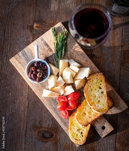 Flat lay food photograph of bruschetti with cheese, cherry tomatoes, olives and red wine over a wooden board on brown background photo