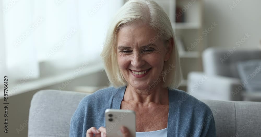 Older generation using modern wireless technology. Attractive smiling mature female sits resting on couch holding smartphone, chatting on-line, spend leisure on internet, watch funny videos, close up Older generation using modern wireless technology. Attractive smiling mature female sits resting on couch holding smartphone, chatting on-line, spend leisure on internet, watch funny videos, close up