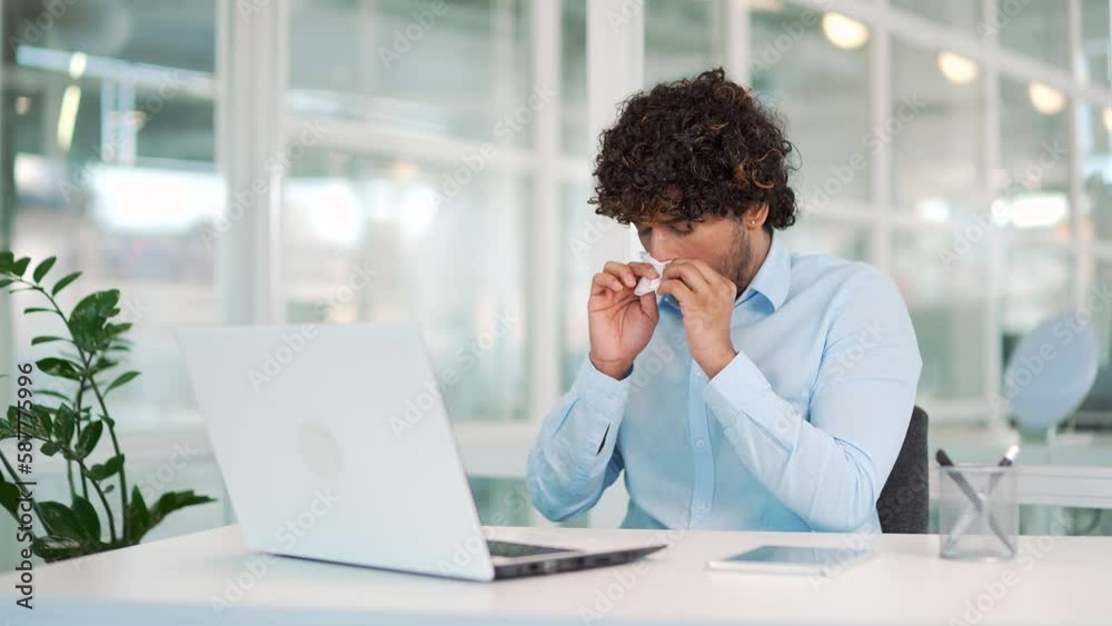 Sick young man suffering from seasonal allergies working on a laptop while sitting at a workplace at a desk in a modern office. The allergic male sneezes and wipes his nose with a handkerchief Sick young man suffering from seasonal allergies working on a laptop while sitting at a workplace at a desk in a modern office. The allergic male sneezes and wipes his nose with a handkerchief