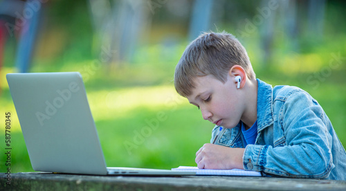 Student with a laptop and a notebook in headphones writing behind a table in a park. Boy is writing down notes outdoors. Scholar studying online with a computer. Boy on online lessons outside..