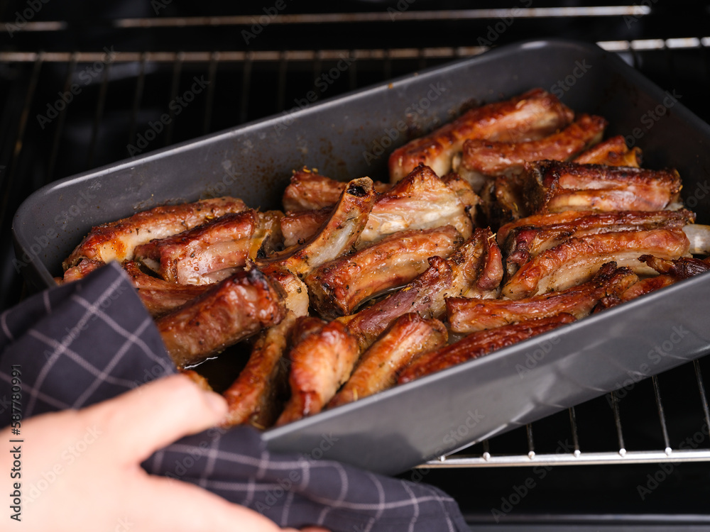 Woman taking black baking tray with baked pork ribs out of an oven