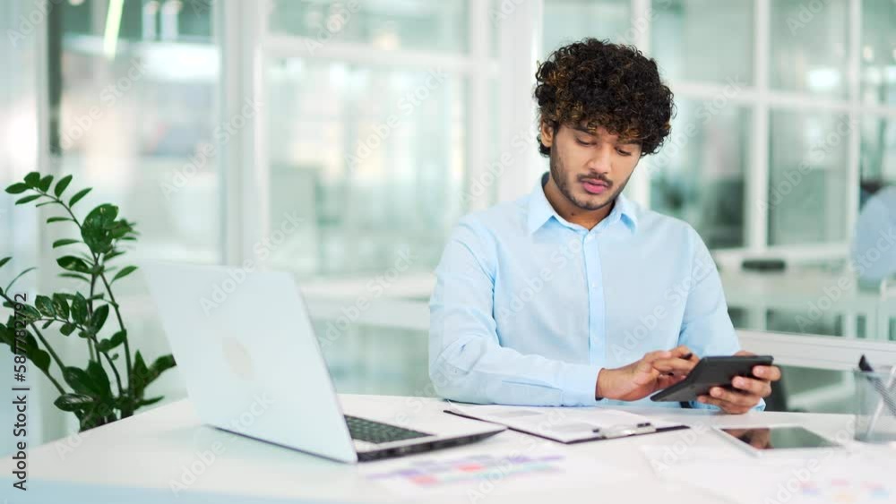 Concentrated accountant calculates production costs on a calculator while sitting at a workplace at a desk in modern office. Serious financier in a shirt writes down data in a notebook using a laptop Concentrated accountant calculates production costs on a calculator while sitting at a workplace at a desk in modern office. Serious financier in a shirt writes down data in a notebook using a laptop
