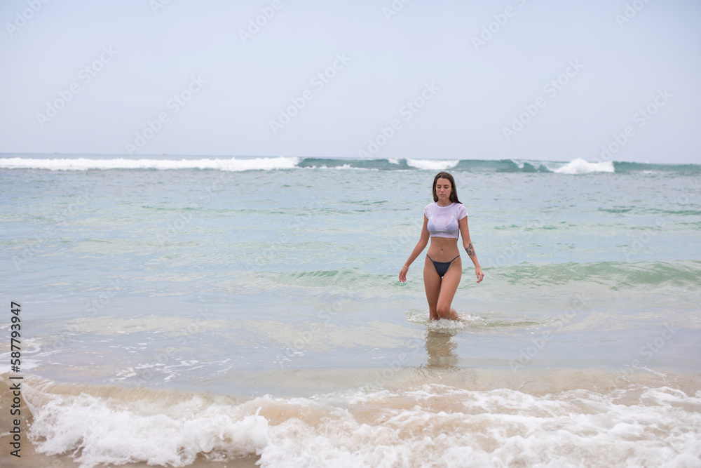Beautiful young woman in a white cropped t-shirt comes out of the ocean. Beautiful young woman in a white cropped t-shirt comes out of the ocean.