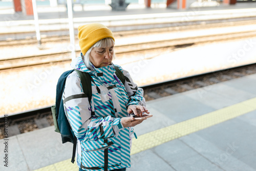 Old happy woman with backpack in yellow hat waiting train on station platform and using smart phone on urban background. Railroad transport concept