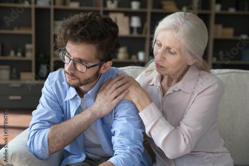 Concerned senior mother talking to frustrated depressed adult son, giving comfort, family support, touching shoulders, helping to cope with problems, loss, sitting close on couch at home