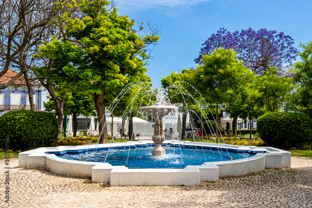 The Fonte do Jardim 1 de Dezembro fountain surrounded by lush vegetation stands in the idyllic Jardim Primeiro de Dezembro park in Portimão with a coffee bar nestled under a purple jacaranda tree.