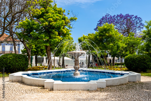 The Fonte do Jardim 1 de Dezembro fountain surrounded by lush vegetation stands in the idyllic Jardim Primeiro de Dezembro park in Portimão with a coffee bar nestled under a purple jacaranda tree.