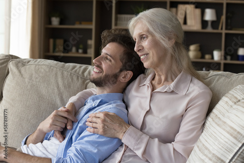 Cheerful elder grandma hugging young adult grandson guy on home couch, enjoying family meeting, leisure time, holding hands, smiling, dreaming on future plans, looking away