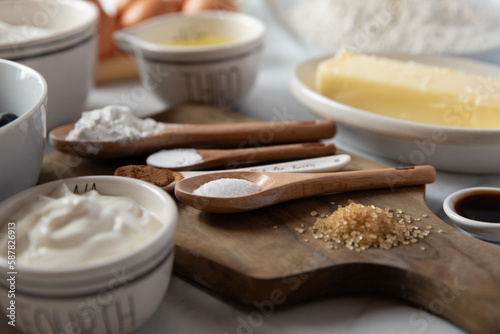 Baking ingredients in measuring spoons and bowls sit on a white marble surface and wooden cutting board.