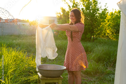 Young woman hanging laundry outdoors. Beautiful girl in red polkadot dress. working in countryside. Cute brunette girl in dress washing white clothes in metal basin in backyard. village girl 