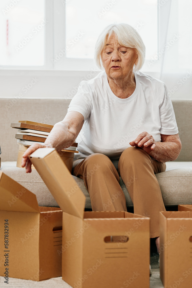elderly woman sits on a sofa at home with boxes. collecting things with memories albums with photos and photo frames moving to a new place cleaning things and a happy smile. Lifestyle retirement. elderly woman sits on a sofa at home with boxes. collecting things with memories albums with photos and photo frames moving to a new place cleaning things and a happy smile. Lifestyle retirement.