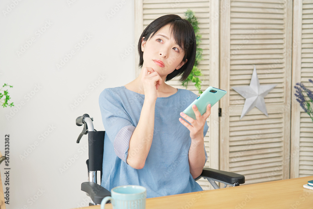 リビングでスマートフォンを使いながら考える車椅子に乗った女性 リビングでスマートフォンを使いながら考える車椅子に乗った女性