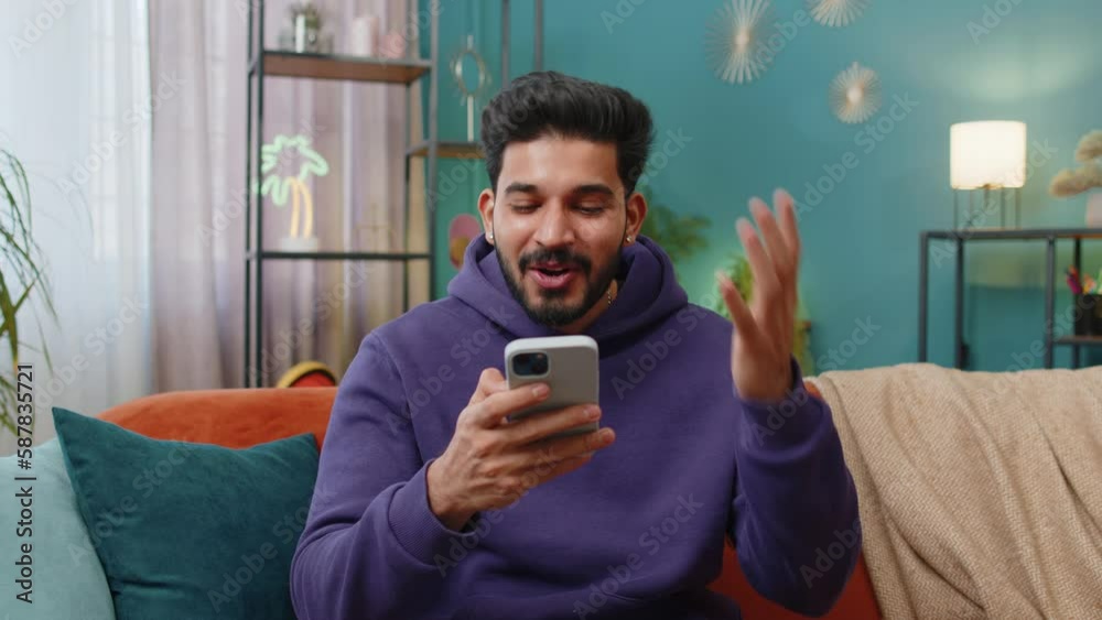 Happy indian man use mobile smartphone typing browsing say Wow yes found out great big win good news celebrate lottery jackpot doing winner gesture. Hindu young guy at home in room sitting on sofa Happy indian man use mobile smartphone typing browsing say Wow yes found out great big win good news celebrate lottery jackpot doing winner gesture. Hindu young guy at home in room sitting on sofa