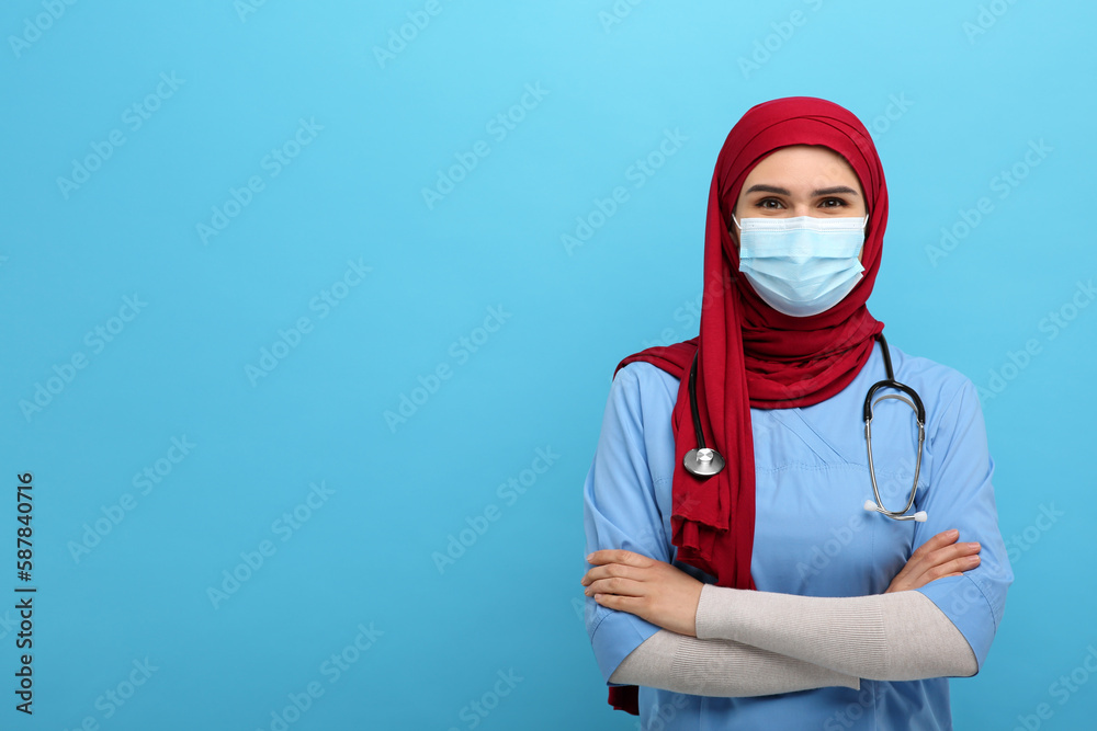 Muslim woman wearing hijab, medical uniform and protective mask on light blue background, space for text