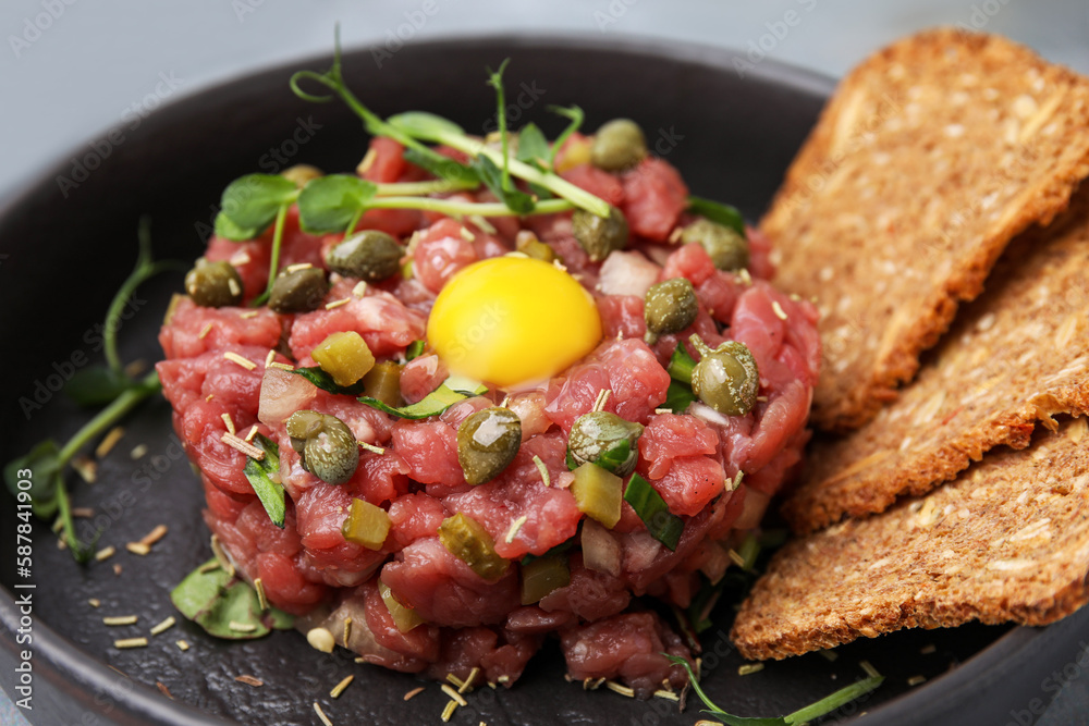 Tasty beef steak tartare served with yolk, capers and bread in serving pan, closeup