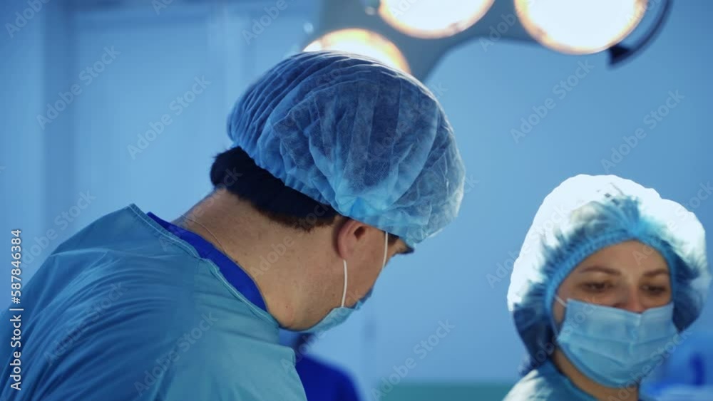 Mid-aged doctor in mask and cap stands under the lamp in surgery room. Nurse brings the materials for procedure. Mid-aged doctor in mask and cap stands under the lamp in surgery room. Nurse brings the materials for procedure.