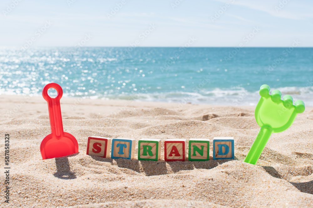 Strand (German Beach) letters on cubes in the sand and ocean in the background. Copy space. Vacations background. Kids sand toys. Strand (German Beach) letters on cubes in the sand and ocean in the background. Copy space. Vacations background. Kids sand toys.
