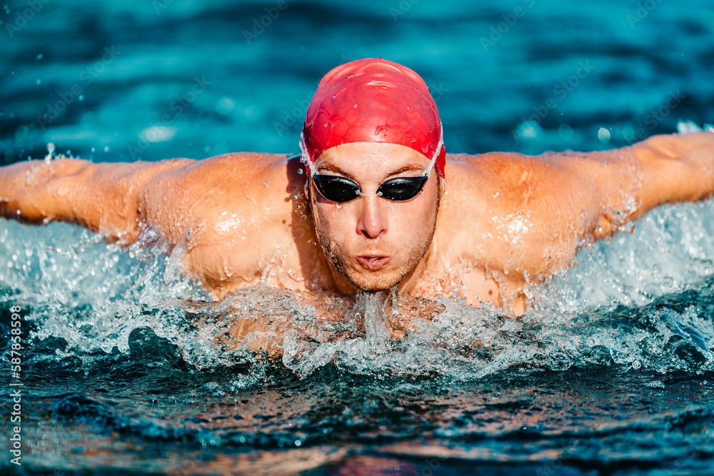Swimming man. Male Swimmer athlete doing butterfly swim stroke. Male sport fitness man wearing swimming goggles and swim cap training hard in outdoor pool. Professional sports fitness model
