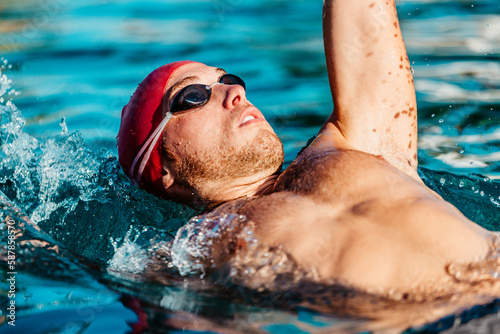 Backstroke Swimming. Man swimming back crawl. Male swimmer doing crawl back stroke in pool wearing swimming goggles and swim cap. Sport, fitness and healthy lifestyle