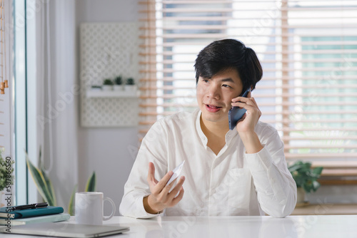 A millennial Asian male freelancer is talking on his smartphone while working remotely from home office with his laptop.