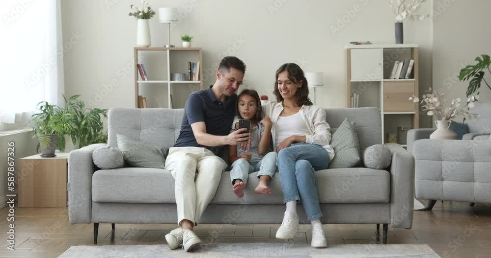 Hispanic couple and child look at smartphone screen, gesturing, grimacing, laughing enjoy new amusing mobile application sit on sofa at home, watch videos, enjoy on-line fun on weekend use modern tech Hispanic couple and child look at smartphone screen, gesturing, grimacing, laughing enjoy new amusing mobile application sit on sofa at home, watch videos, enjoy on-line fun on weekend use modern tech