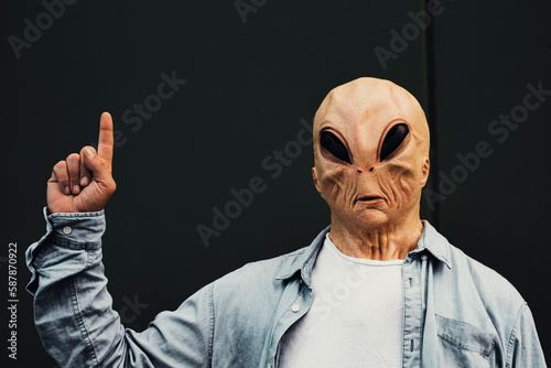 Ufo alien portrait standing against a black wall background and pointing finger hand in talking posture gesture. Copy write and your text here. Commercial advice with person masked and rare