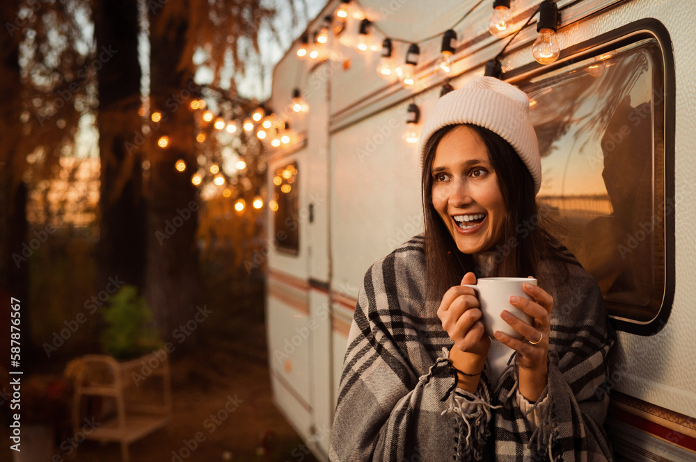 Caucasian woman in a knitted hat wrapped in a plaid and drinks a warming drink outdoors. Travel in a motor home in the fall.