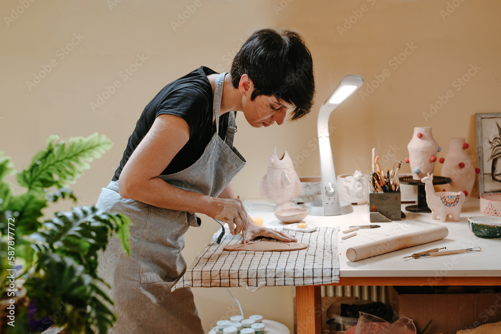 Young woman potter carves a plate from clay in her workshop.