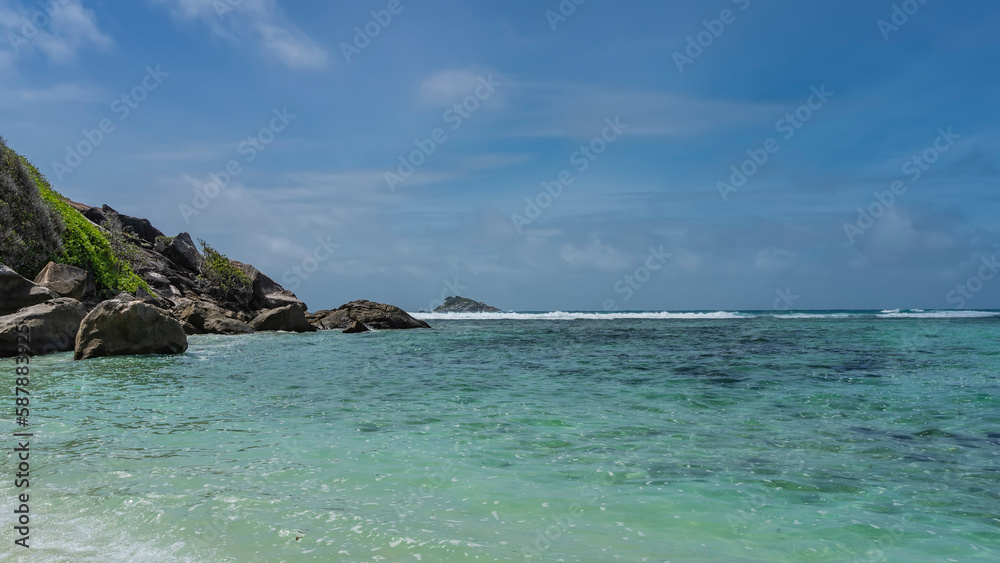 Tropical idyll. Calm turquoise ocean and blue sky with clouds. The foam of waves over the reef in the distance.  Green vegetation on the rocky slope of the island. Seychelles. Moyenne Island. 