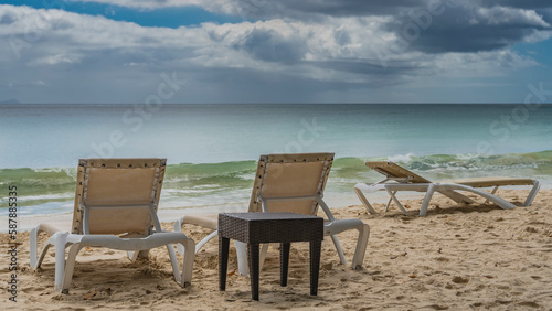 Sun loungers and a wicker table stand on the sandy beach. The waves of the turquoise ocean roll ashore. Clouds in the blue sky. Seychelles. Mahe. Beau Vallon © Вера 