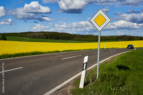 Traffic sign on the side of the road. Yellow rapeseed (Brassica napus) field behind the roadway. Green forest in front of blue cloudy sky. Driving vehicle.