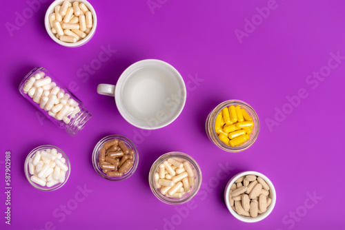 Organic food supplements in small jars and a cup of water from above on violet background. Vitamins and minerals in capsules.