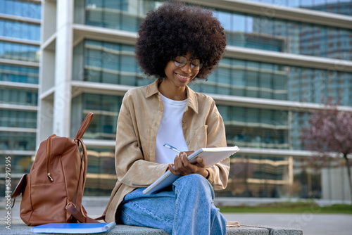 Happy young black African American woman university student doing homework, learning and taking notes in notepad sitting outside uni campus area on fresh air, near trees and buildings and smiling.