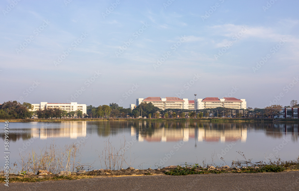Panoramic view of buildings near trees next to the shore of a large pond with water reflections.
