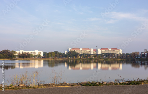 Panoramic view of buildings near trees next to the shore of a large pond with water reflections. © kaentian
