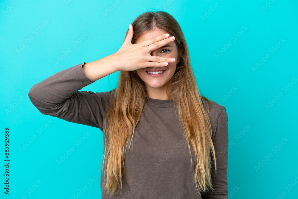Young blonde woman isolated on blue background covering eyes by hands and smiling