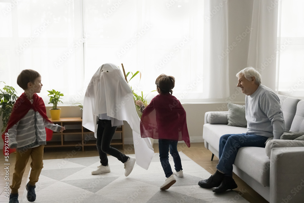 Fototapeta premium Happy senior grandfather watching three boys in homemade super hero costumes chasing each other, running, playing role active game. Grandpa enjoying meeting with grandsons