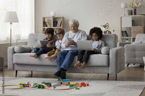 Older grandpa watching multiethnic grandkids, resting on couch with children, using mobile phone. Diverse kids holding digital gadgets, sitting on sofa near granddad engaged in online addiction