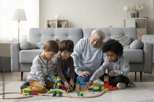 Elderly grandpa watching three multiethnic little grandsons playing game with toy construction blocks on warm floor, building city towers model together, enjoying family playtime, teamwork