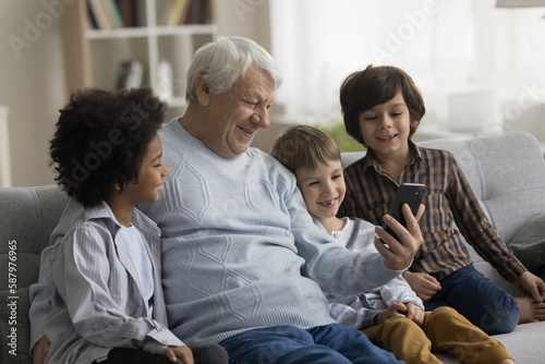 Cheerful grandpa and diverse little grandsons talking on video conference call on smartphones, sitting on couch, using cellphone for online communication, laughing, having fun